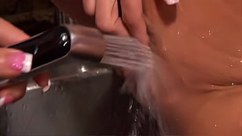 Sexy lesbians stimulate their pussies with a stream of water and fuck themselves with a strap on in a standing position from behind