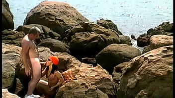 A mature man fucks an exotic babe wearing only a cap and sunglasses on the seaside stones